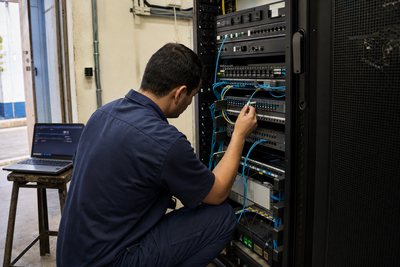 Técnico ajoelhado conecta cabeamento em um rack de rede aberto, com notebook de diagnóstico ao lado